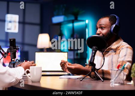 Un jeune homme noir avec un carnet donne des conseils en ligne en utilisant un équipement de podcast. A la table, un vlogger afro-américain parle dans le microphone, enregistrant un épisode à la maison. Banque D'Images