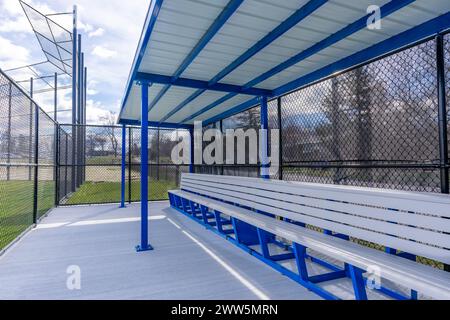 Vue de la dugout typique de baseball de lycée non descript avec sol en béton, clôture à maillons de chaîne et abat-jour bleu sur le toit. Banque D'Images