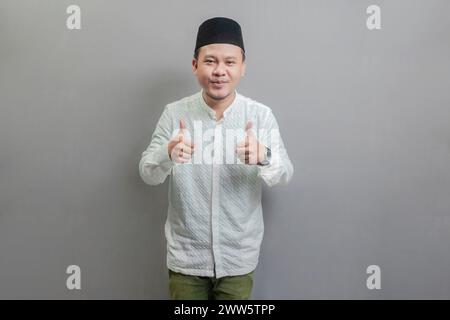 Heureux homme musulman asiatique avec la main, les pouces souriants et heureux pour le succès. Geste gagnant portant une chemise koko et peci avec des nuances de mon jeûne Banque D'Images