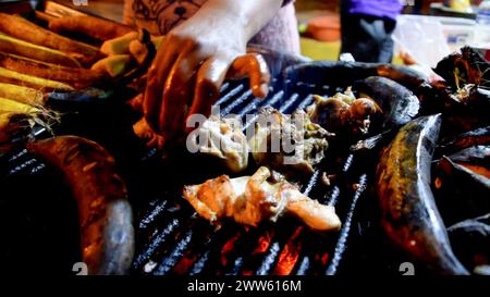 Diverses viandes et légumes cuisent sur un barbecue fumé dans un cadre animé de marché nocturne Banque D'Images