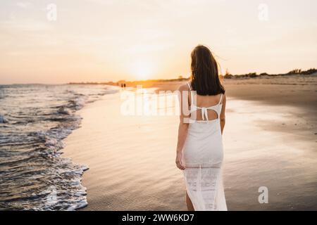belle femme marchant le long du rivage de la plage avec la lumière du coucher du soleil habillée en blanc Banque D'Images