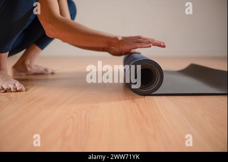 Gros plan des mains d'une femme roulant un tapis de yoga dans une pièce avec un plancher de dais. Banque D'Images