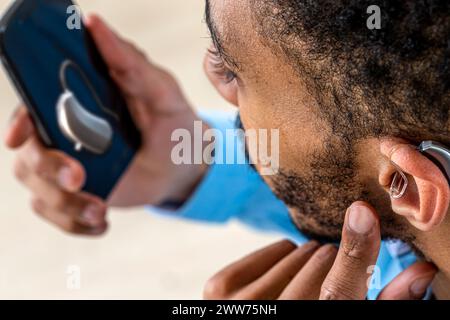 Jeune homme ajustant l'intensité du volume de l'aide auditive avec le smartphone. Banque D'Images