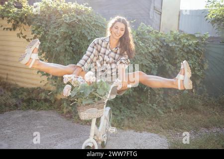 Une jeune femme attirante en vêtements décontractés est assise sur un vélo dans le jardin. Une charmante fille aux cheveux bouclés sourit dehors dans le soleil lumineux Banque D'Images