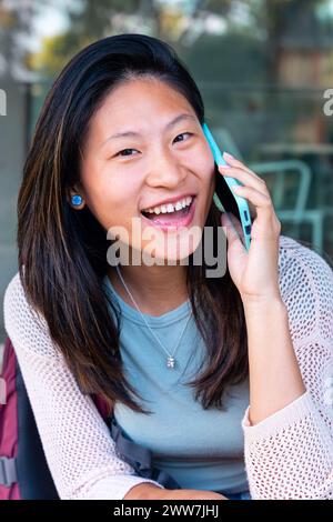 Portrait vertical jeune femme asiatique souriante assise parlant sur smartphone regardant la caméra à l'extérieur. Banque D'Images