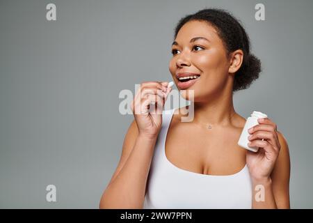 heureuse femme afro-américaine prenant des suppléments sous forme de pilule sur fond gris, plan diététique Banque D'Images