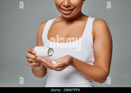 femme afro-américaine coupée versant des pilules de la bouteille dans la main sur fond gris, plan diététique Banque D'Images