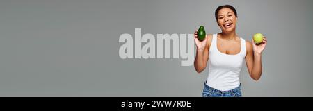 Femme afro-américaine rayonnante avec pomme et avocat favorisant une nutrition équilibrée sur bannière grise Banque D'Images