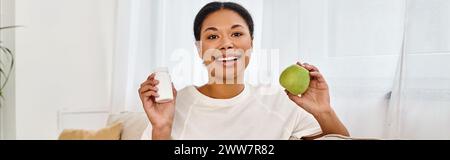 diététiste afro-américain tenant pomme et suppléments et souriant dans le salon, bannière Banque D'Images