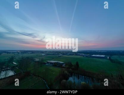 Une vue aérienne capture les vastes serres industrielles alors que le jour cède à la nuit. Le coucher de soleil jette une lueur chaude sur le paysage, créant un équilibre frappant entre les éléments industriels et la sérénité naturelle de la campagne. Coucher de soleil sur l'harmonie industrielle : une fin tranquille à une journée dans l'agriculture. Photo de haute qualité Banque D'Images