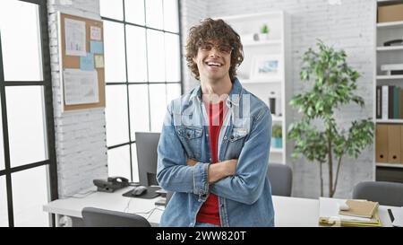 Jeune homme hispanique souriant avec barbe et lunettes en veste denim croisant les bras dans un bureau moderne. Banque D'Images