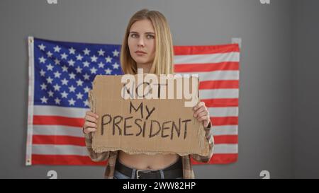 Une femme blonde caucasienne tient une pancarte de protestation avec un drapeau américain en toile de fond véhiculant une dissidence politique Banque D'Images