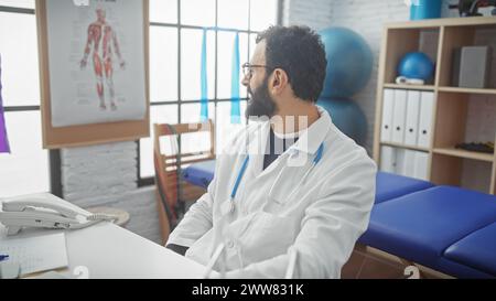 Un bel homme barbu vêtu d'un manteau blanc s'assoit soigneusement dans la salle de consultation d'une clinique médicale. Banque D'Images