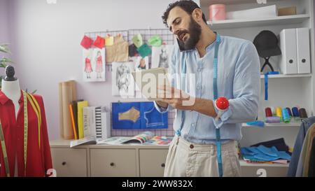 Un tailleur barbu dans une chemise bleue multitâche, parlant au téléphone tout en examinant des notes dans un atelier coloré avec des mannequins et des tissus. Banque D'Images