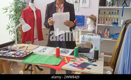 Femme africaine tailleur examinant des motifs dans un atelier coloré avec des tissus et une machine à coudre Banque D'Images