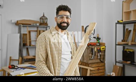 Un jeune homme hispanique joyeux avec une barbe se tient confiant dans un atelier de menuiserie tenant du bois. Banque D'Images