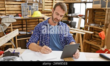 Artisan barbu en chemise bleue à l'aide de la tablette et de l'écriture dans un atelier de menuiserie lumineux Banque D'Images