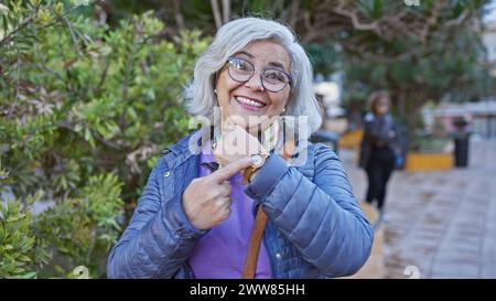 Une femme mature souriante avec des lunettes et des cheveux gris pointe à sa montre dans un cadre de parc extérieur vert. Banque D'Images