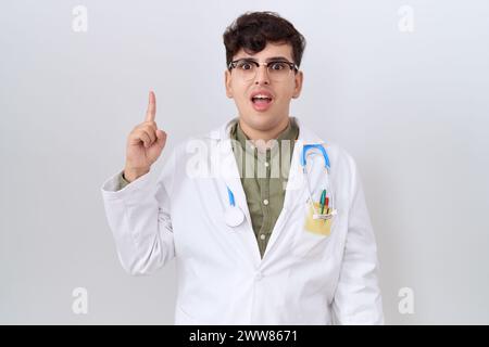 Jeune homme non binaire portant l'uniforme du médecin et le stéthoscope pointant le doigt vers le haut avec l'idée réussie. sorti et heureux. numéro un. Banque D'Images