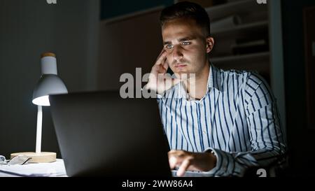 Bel homme hispanique travaillant tard dans un bureau faiblement éclairé, concentré sur l'écran d'un ordinateur portable. Banque D'Images