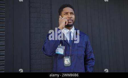 Un homme afro-américain concentré en uniforme parlant sur un téléphone avec un badge visible, debout sur un fond urbain. Banque D'Images