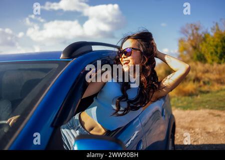 Un jeune asiatique se penche par la fenêtre de la voiture et rit. Une femme coréenne souriante voyage et s'amuse sur la route. Une fille attrayante aime le trajet et n Banque D'Images