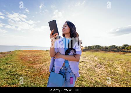 Une jeune fille asiatique attrayante avec de longs cheveux ondulés tient un téléphone dans une clairière. Une charmante femme coréenne dans des vêtements décontractés rit et utilise un gadg mobile Banque D'Images