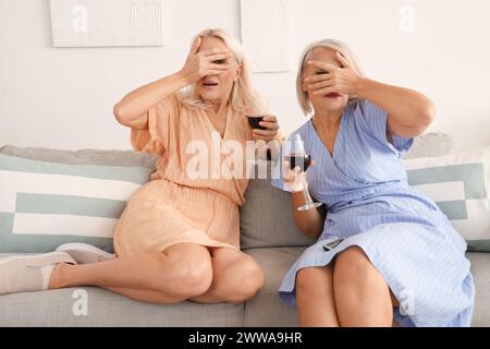 Des amies matures choquées avec des verres de vin regardant la télévision à la maison Banque D'Images