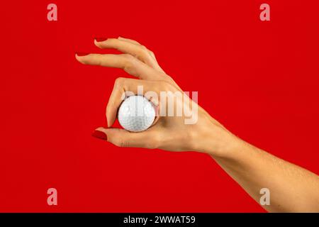 Main de femme anonyme de culture avec des ongles peints et des doigts montrant une petite balle de golf blanche sur fond rouge dans le studio Banque D'Images