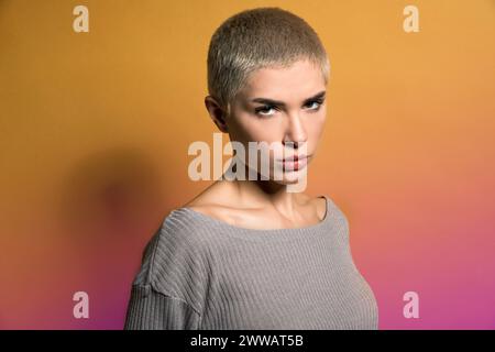 Modèle jeune femme sérieux avec des cheveux blonds courts dans des vêtements décontractés regardant la caméra avec attention contre fond dégradé de deux couleurs Banque D'Images