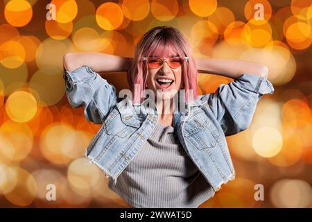Joyeuse dame élégante en lunettes de soleil et veste en denim recadrée riant avec les mains derrière la tête sur fond bokeh orange Banque D'Images