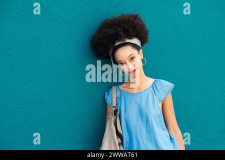 Portrait de jeune femme marocaine souriante dans le maquillage avec bandeau afro cheveux regardant la caméra tout en se tenant debout avec sac à main sur fond bleu en d. Banque D'Images