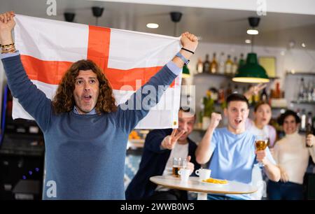 Compagnie enthousiaste de jeunes fans de sport adultes brandissant le drapeau de l'Angleterre et soutenant l'équipe nationale avec de la bière dans le pub Banque D'Images