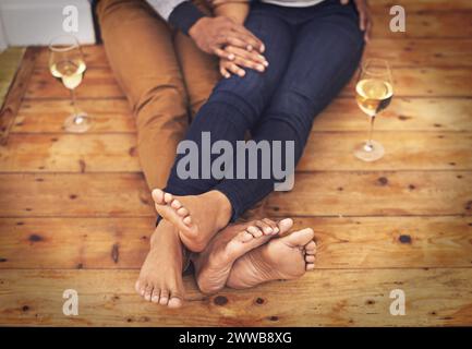 Vin, pieds et couple se relaxant ensemble sur plancher en bois pour une date romantique à la maison moderne. Amour, champagne et gros plan des jambes de l'homme et de la femme avec Banque D'Images