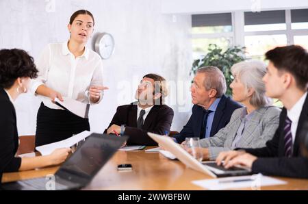 Intelligent jeune femme faisant rapport tandis que les employés de bureau d'âges différents utilisant des cahiers à la table dans le bureau Banque D'Images