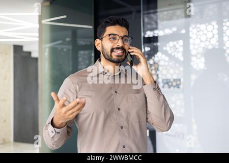 Un homme parle sur son téléphone portable dans une pièce avec un mur de verre. Il porte une chemise marron et des lunettes Banque D'Images