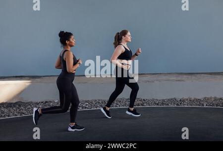 Deux femmes avec différents types de corps courir à l'extérieur. Vue latérale de deux jeunes femelles qui font du jogging sur un mur gris. Banque D'Images