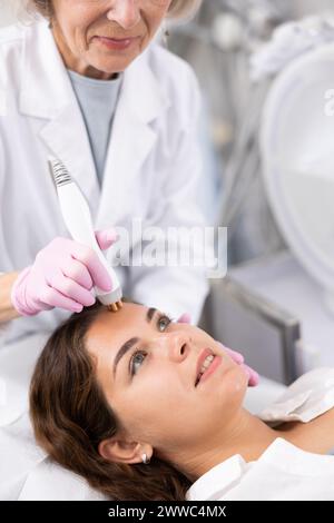 Médecin esthéticien âgé faisant la procédure de levage de RF pour la pose de visage de femme impeccable dans le salon de beauté. Cosmétologie matérielle. Banque D'Images