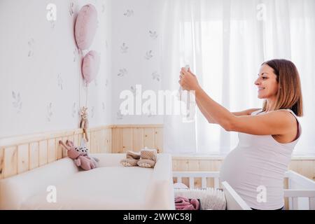 Femme enceinte souriante arrangeant les vêtements de bébé dans la chambre de la crèche Banque D'Images