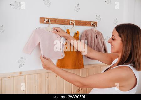 Femme enceinte souriante accrochant des vêtements de bébé sur le mur à la maison Banque D'Images