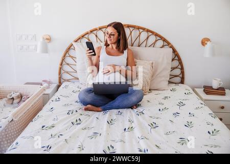 Freelance enceinte utilisant un téléphone intelligent sur le lit au bureau à domicile Banque D'Images