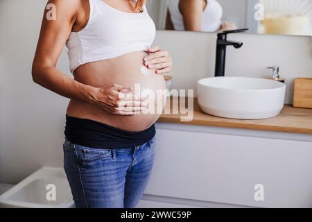 Femme enceinte appliquant la crème sur le ventre dans la salle de bain à la maison Banque D'Images