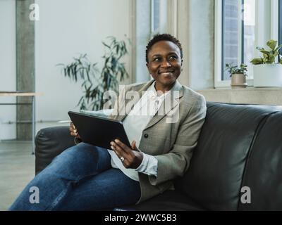 Femme d'affaires souriante portant blazer assis sur le canapé avec tablette PC dans le bureau Banque D'Images