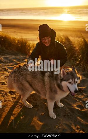 Jeune femme souriante caressant chien Husky sur le sable à la plage Banque D'Images