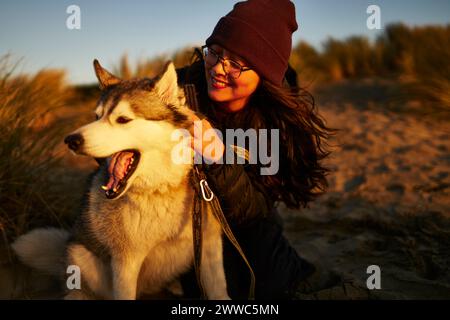 Femme souriante portant un chapeau en tricot et caressant chien Husky au coucher du soleil Banque D'Images