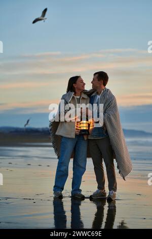 Homme et femme souriant avec lanterne enveloppée dans un châle à la plage de l'océan Banque D'Images