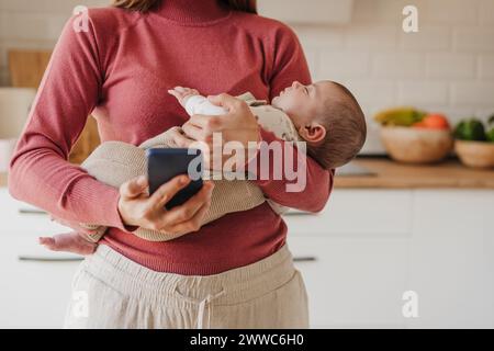 Mère à l'aide d'un téléphone intelligent et transportant la petite fille mignonne dans la cuisine à la maison Banque D'Images