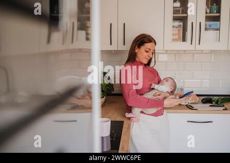 Femme portant la fille bébé endormie et utilisant le téléphone intelligent dans la cuisine Banque D'Images