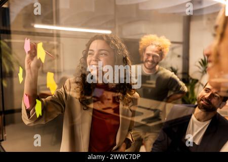 Femme d'affaires souriante faisant séance de brainstorming avec des collègues derrière verre au bureau Banque D'Images