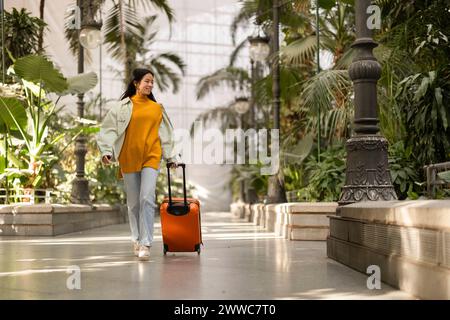 Heureuse jeune femme marchant avec une valise près des plantes Banque D'Images